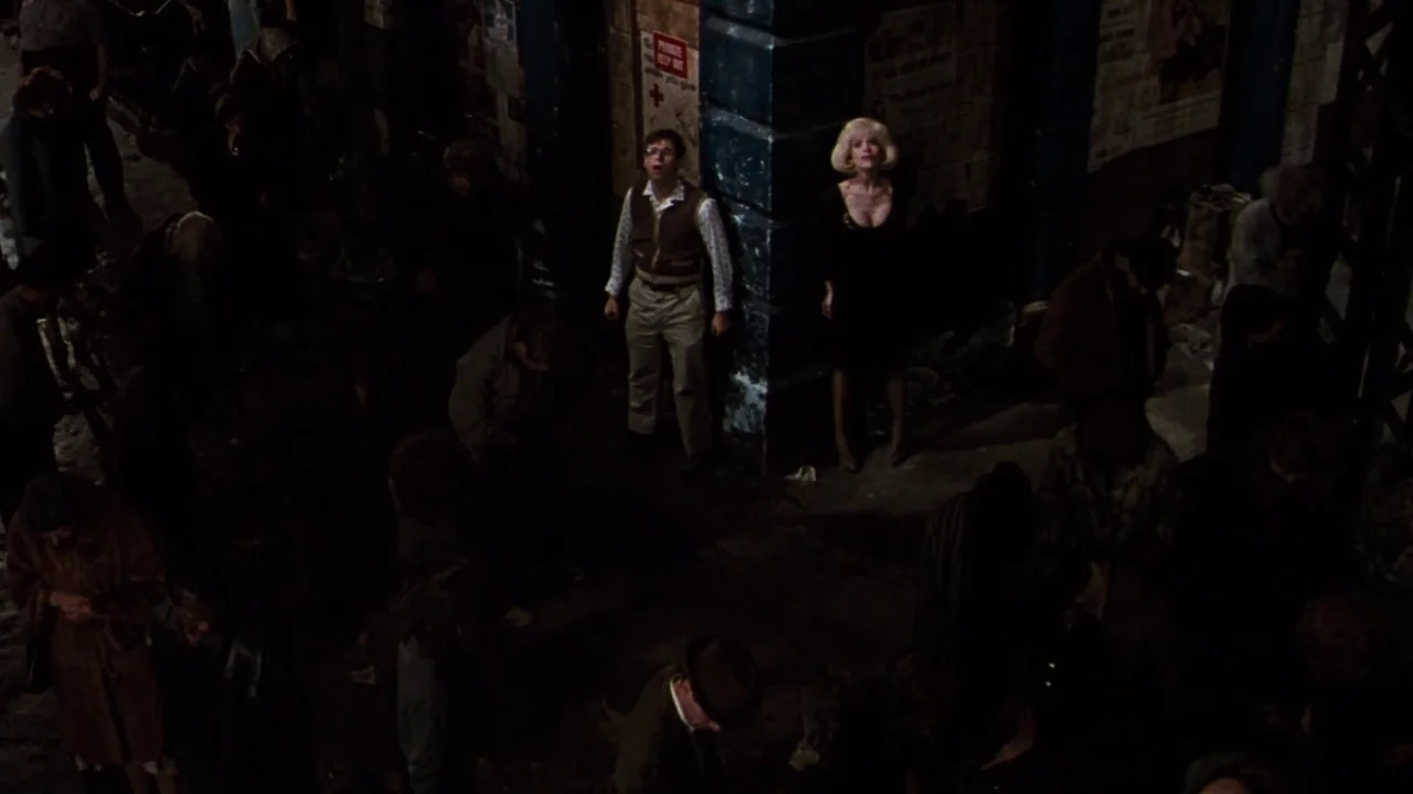 Little Shop of Horrors (1986) movie still: wide — A dark, gritty city street corner at night with posters plastered on brick walls. A man and woman…