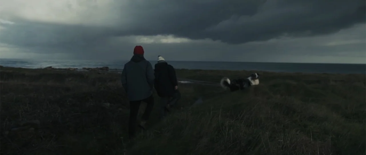 Taking Back the Waves (2017) movie still: wide — A rugged grassy coastline sits under a heavy, dark overcast sky at dusk. Two people in winter…