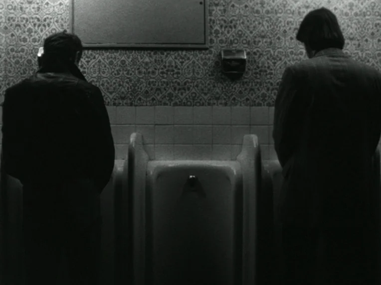 Death and Transfiguration (1983) movie still: medium — A public restroom features patterned wallpaper and white ceramic tiling above the fixtures.…