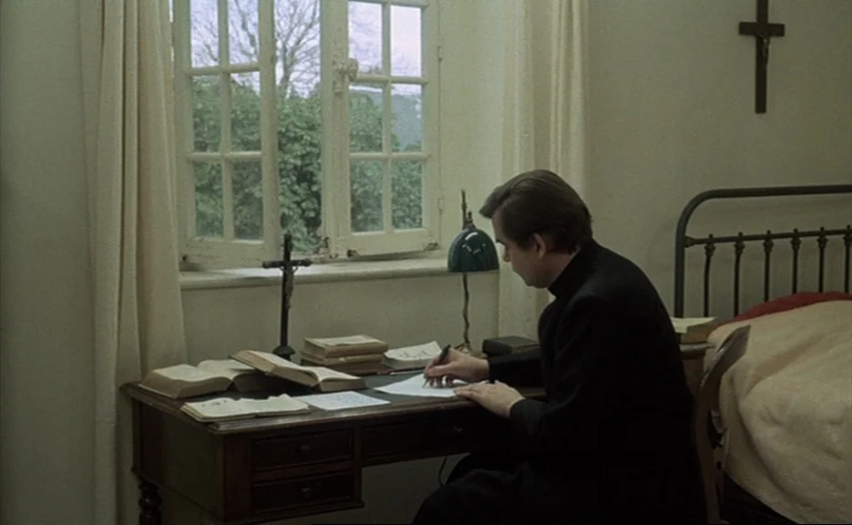 Murmur of the Heart (1971) movie still: medium — A spartan room features a desk by a window with a crucifix on the wall. A priest in a black cassock…