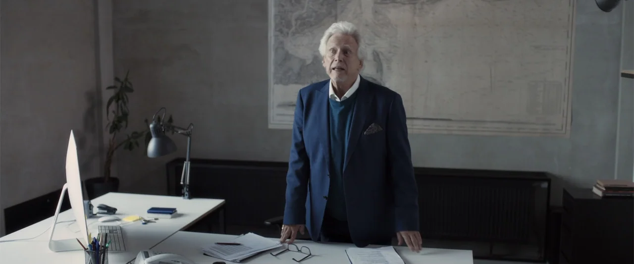 The Good Neighbor (2022) movie still: medium — A minimalist office contains a white desk, a map on the wall, and a plant. An older man stands behind…