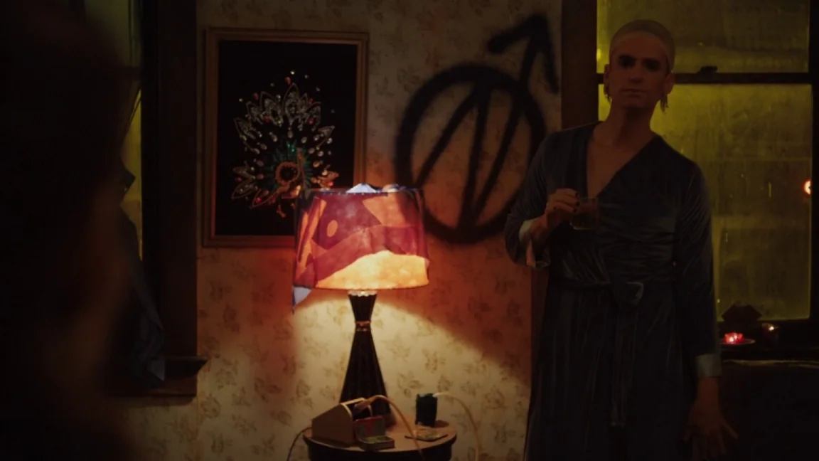 Adult World (2013) movie still: medium — A dimly lit room features floral wallpaper with a large black painted symbol. The person stands near a…