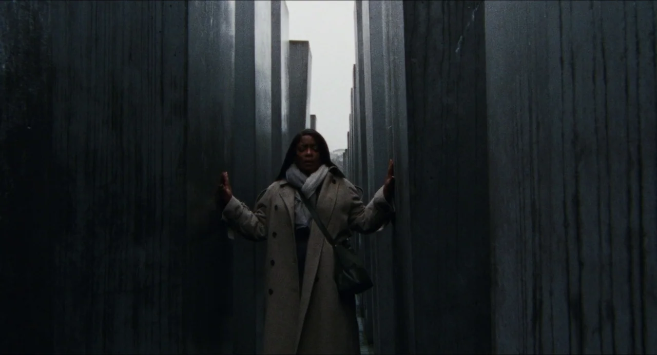 Origin (2023) movie still: fullbody — Rows of tall, dark, rectangular concrete pillars stand in a grid formation outdoors. A woman in a beige coat…
