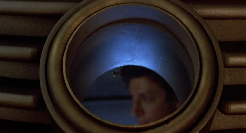 The Fly (1986) movie still: closeup — A dark, metallic industrial structure features a circular window looking into a blue-lit area. A man's face is…