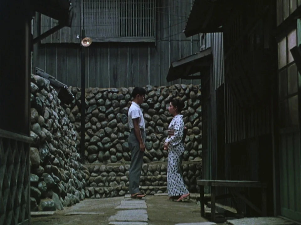 Floating Weeds (1959) movie still: fullbody — A narrow outdoor walkway is enclosed by tall stone retaining walls and wooden structures. A young man…