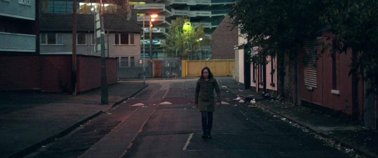The Cured (2017) movie still: fullbody — An empty urban roadway at twilight features low-rise buildings and a distant parking structure. A woman in…