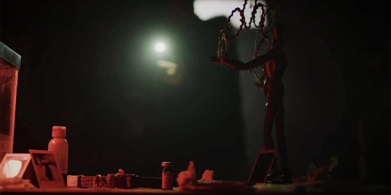 Lobos (2019) movie still: medium — A dark, dimly lit desk holds a small wooden mannequin draped in tangled string. Various personal items and a…