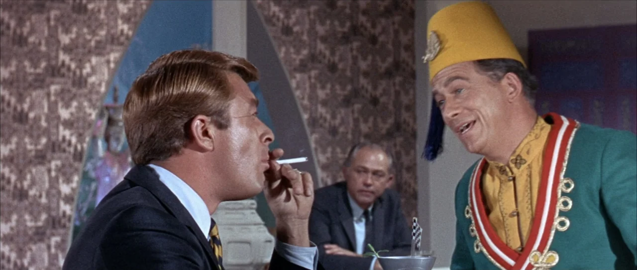 Clambake (1967) movie still: medium — A mid-century dining space with patterned wallpaper features two men sitting at a table. One man in a suit…
