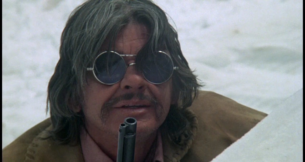 The White Buffalo (1977) movie still: closeup — A snowy, sun-drenched landscape serves as the backdrop for a man hiding in snow. The man wears dark…
