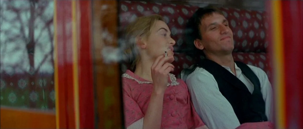 Jude (1996) movie still: medium — A train carriage interior features red patterned upholstery and a large windowed view outside. A woman holds a lit…