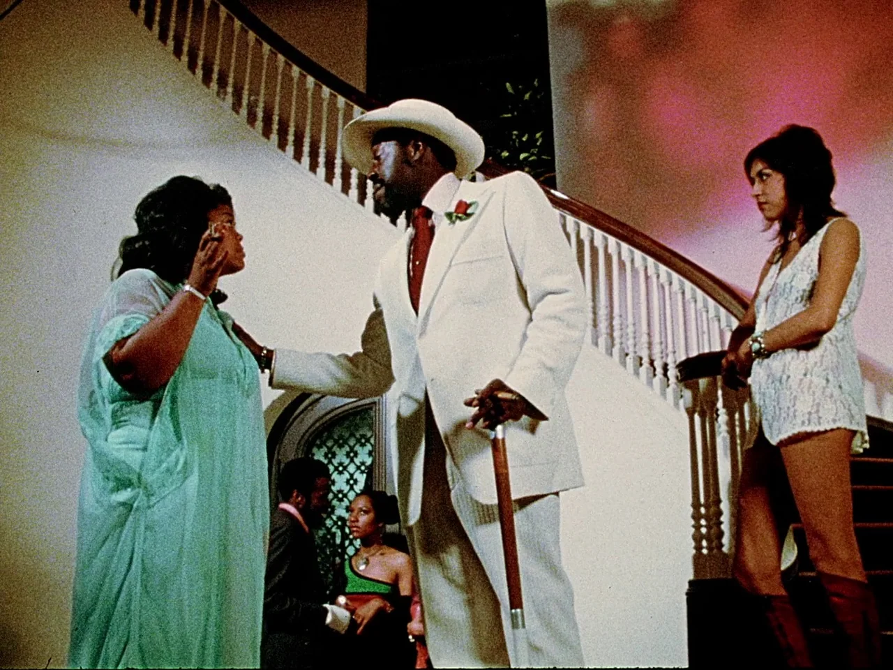 Space is the Place (1974) movie still: fullbody — A grand foyer with a sweeping wooden staircase and a dark-toned decorative screen. A man in a…