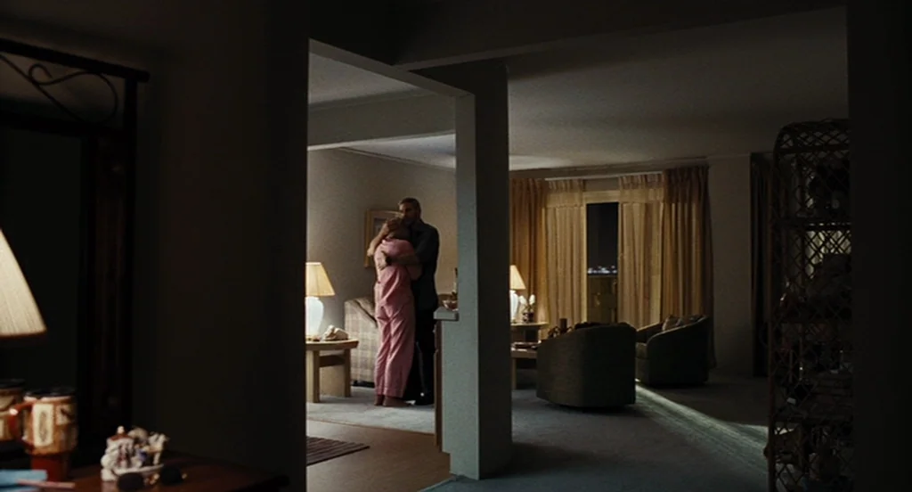 Burn After Reading (2008) movie still: wide — An apartment living room is viewed through a darkened doorway with furniture in shadow. A man and a…