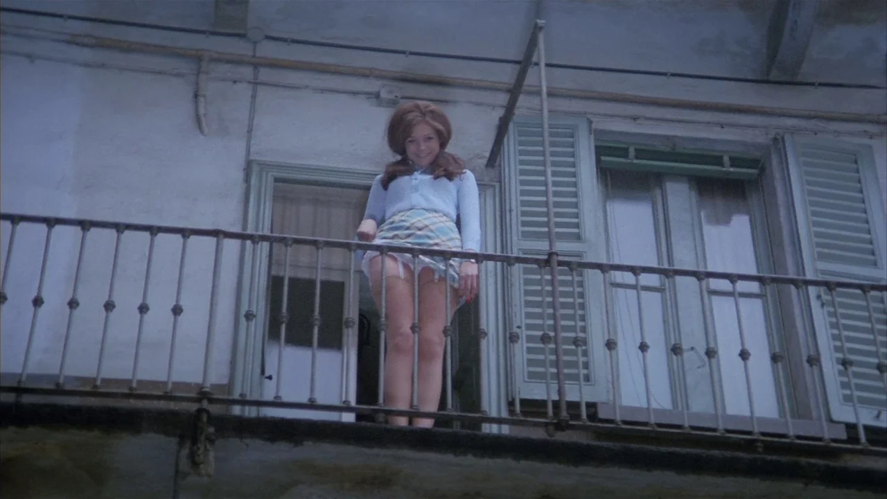 Death Occurred Last Night (1970) movie still: fullbody — An exterior residential building with a weathered white facade features a wrought iron…