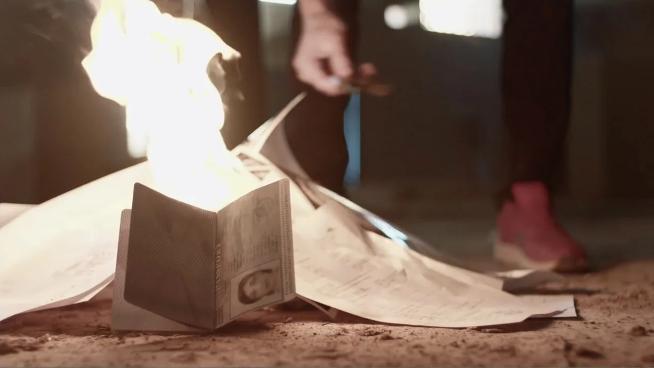 Code of Trust (2019) movie still: closeup — A pile of paper documents and a passport burn on dry dirt ground. Open flames consume the edges of the…