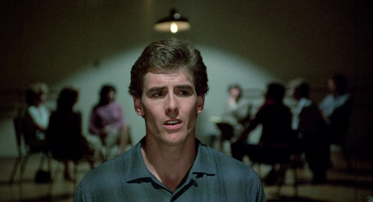 Crimes of Passion (1984) movie still: medium — A sparsely furnished room holds several people sitting in a circle of chairs. A man in the foreground…