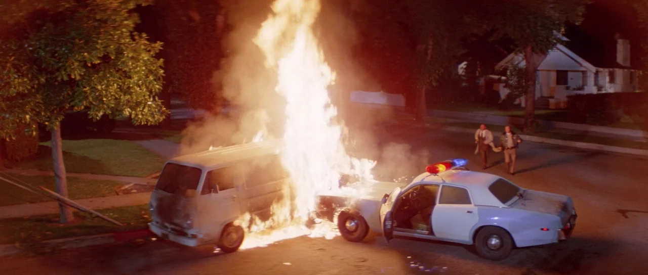Halloween II (1981) movie still: wide — A residential street at night features a van and a patrol car engulfed in flames. Two men walk briskly away…