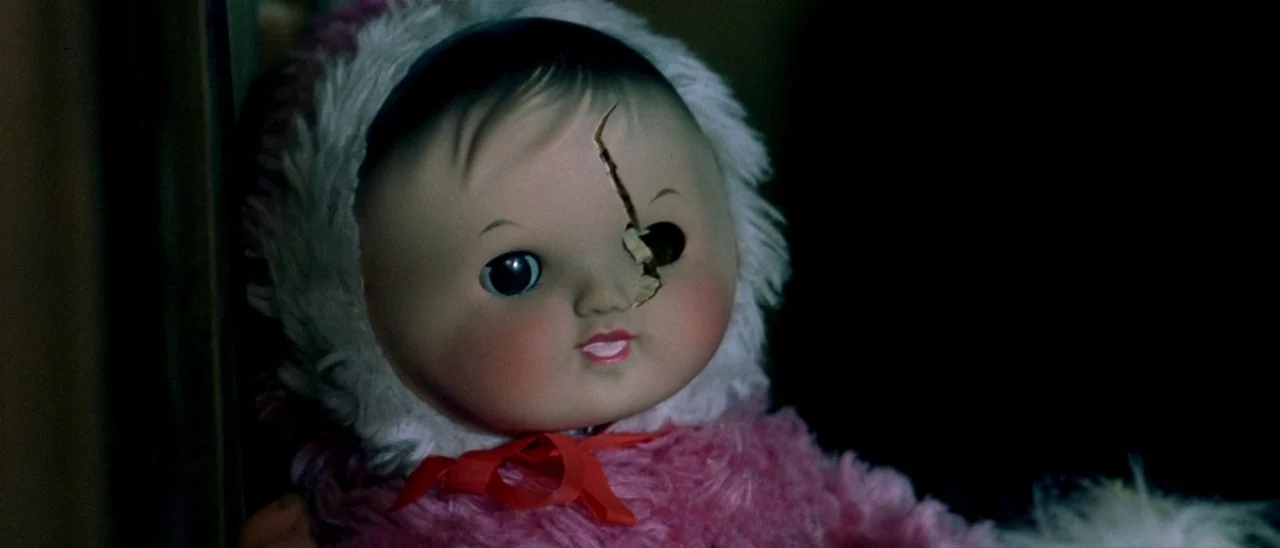 Haute Tension (2003) movie still: closeup — A vintage doll with a cracked face and white fur-trimmed hood occupies the frame. The doll stares…