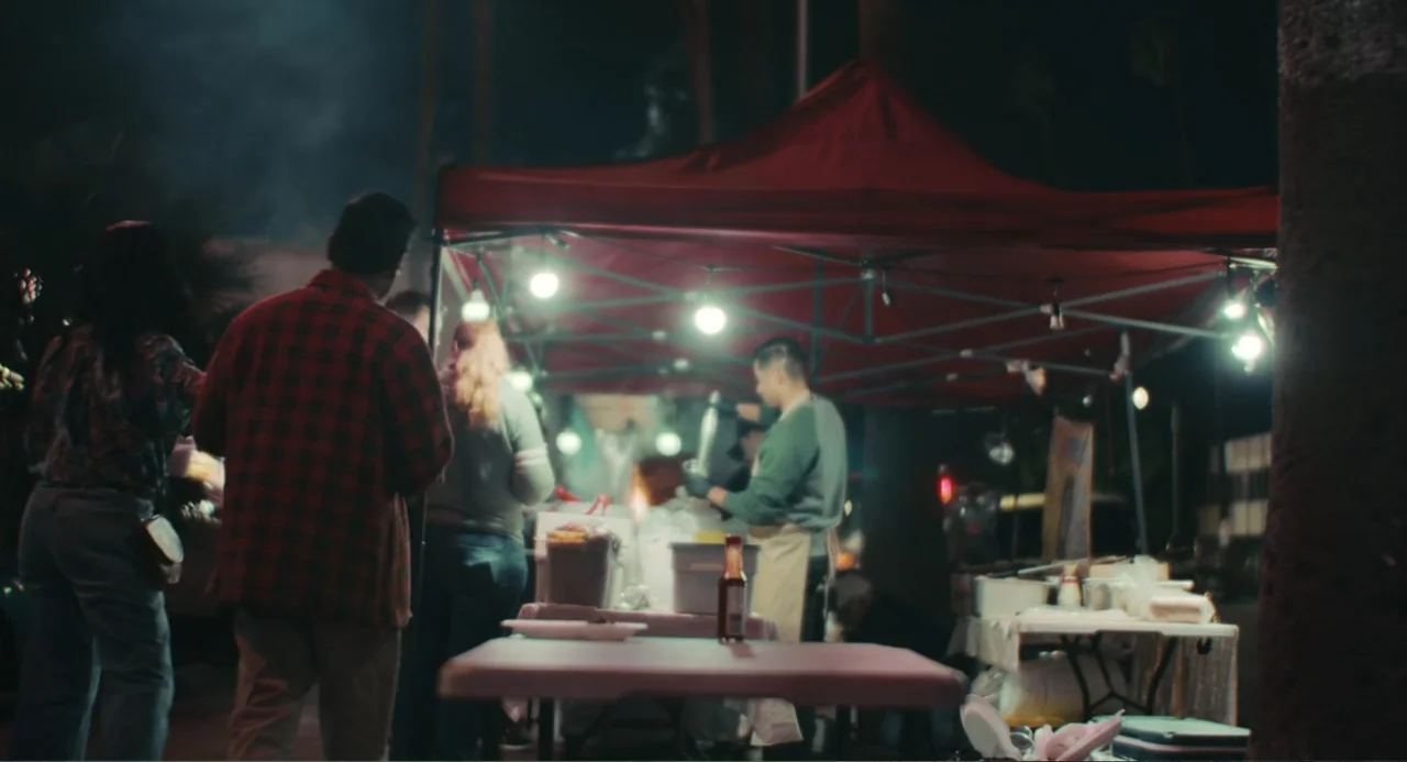 Good Fortune (2025) movie still: medium — A night food stall is covered by a large red pop-up canopy tent. A man stands in line while a vendor works…