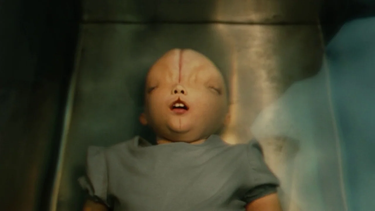 Splice (2009) movie still: closeup — A person lies on a sterile metallic surface inside a cold environment. The subject rests quietly with a…