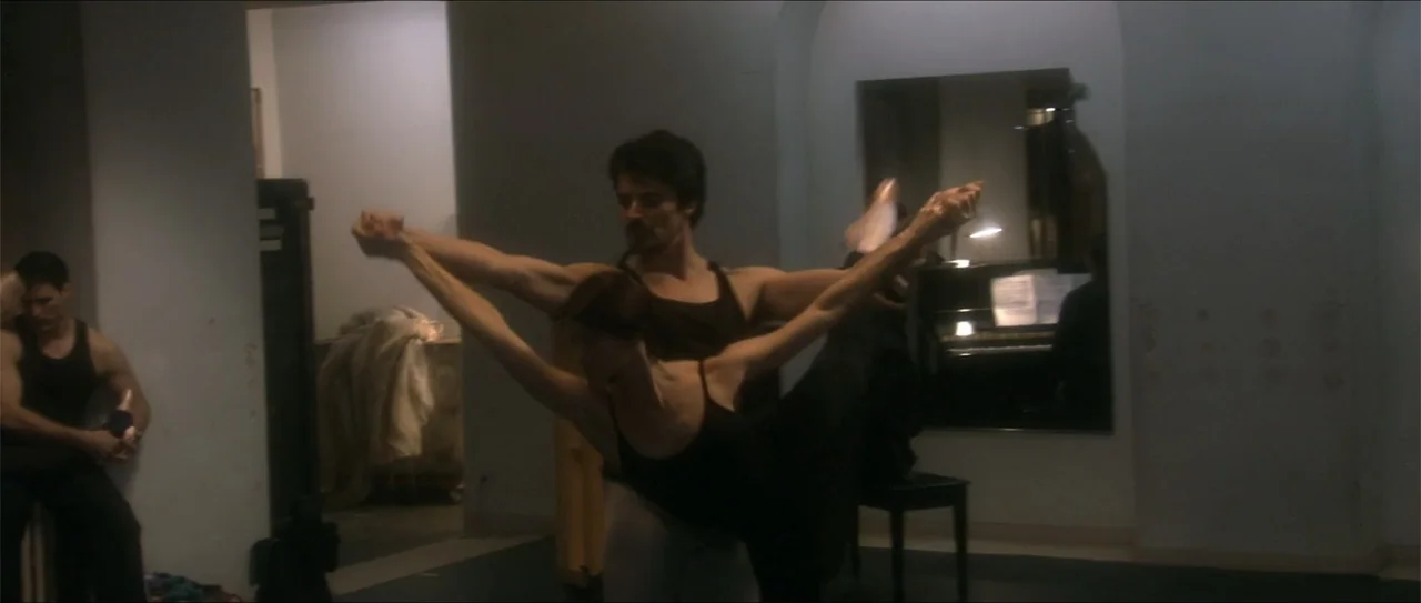 El acto (1989) movie still: medium — A dimly lit dance practice room features a piano against a back wall. Two dancers rehearse an extension…