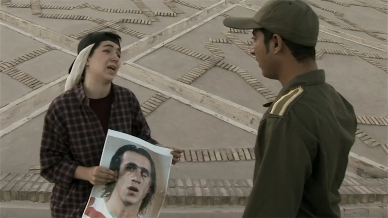 Offside (2015) movie still: medium — A stone-paved plaza with geometric brick patterns serves as the backdrop for two people. A young person in a…