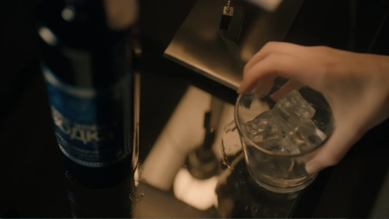 Sightless (2017) movie still: closeup — A dark reflective table surface holds a bottle of vodka and a glass of ice. A person's hand reaches into the…
