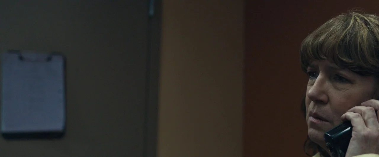 Compliance (2012) movie still: closeup — An office interior with a brown wall and a clipboard on a door. A woman in a yellow shirt holds a black…