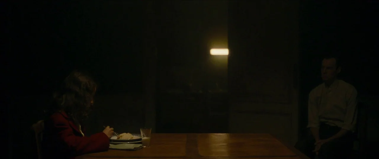 Earwig (2022) movie still: wide — A dark dining room with a wooden table centered in the frame. A young girl eats alone while a man sits silently on…