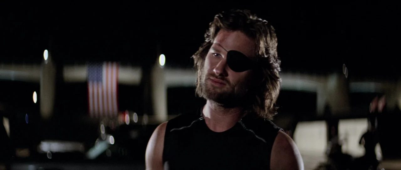 Escape From New York (1981) movie still: medium — An outdoor night scene featuring a man against a dark, blurred structural background. A large…
