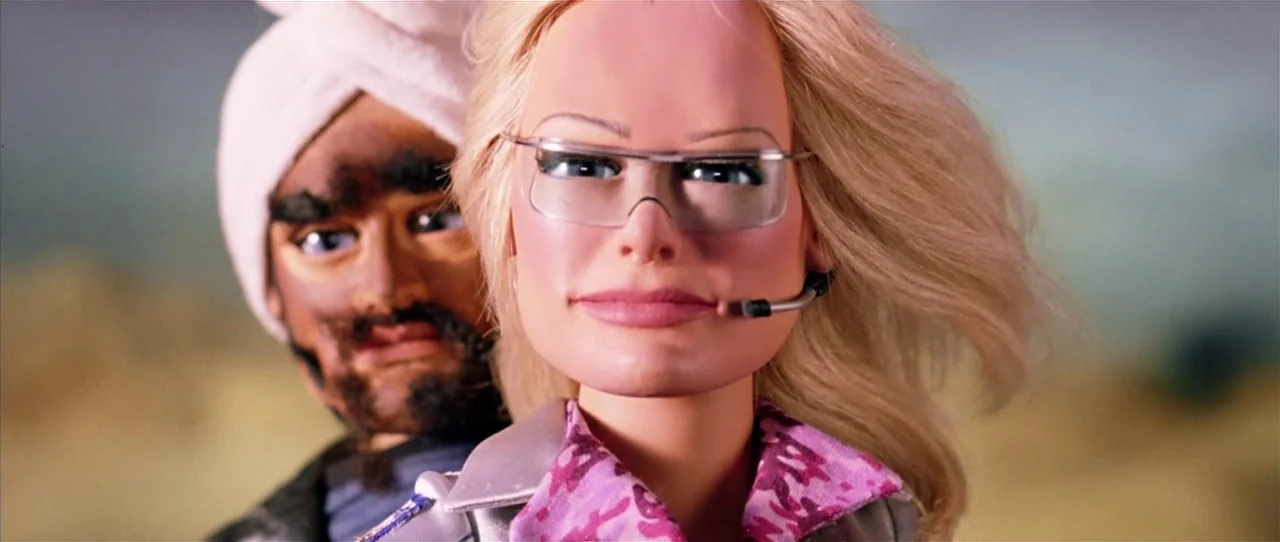 Team America: World Police movie still: medium — Two puppets are positioned against a blurry, light brown outdoor desert landscape. A blonde woman…