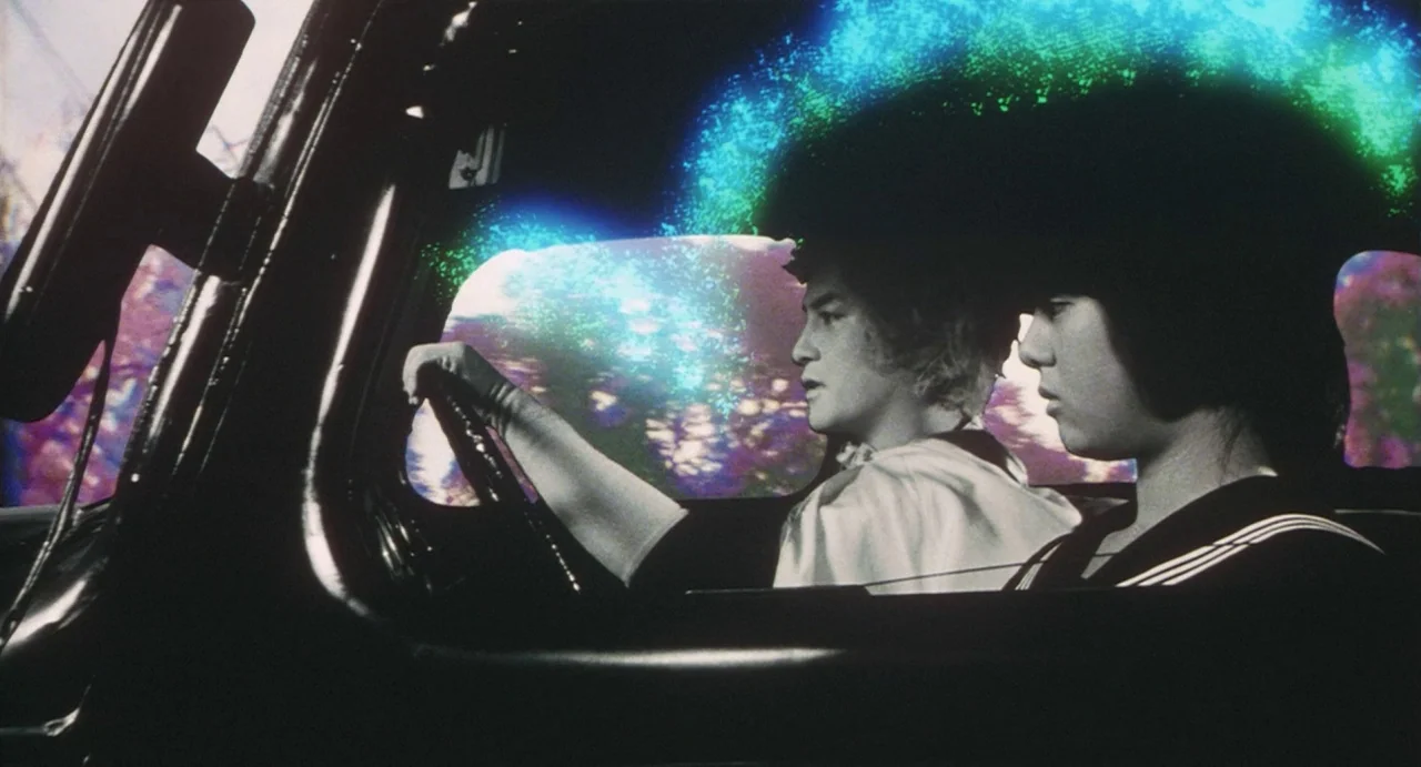 School in the Crosshairs (1981) movie still: medium — Two individuals sit in the front seats of a dark car at night. Neon blue and purple light…