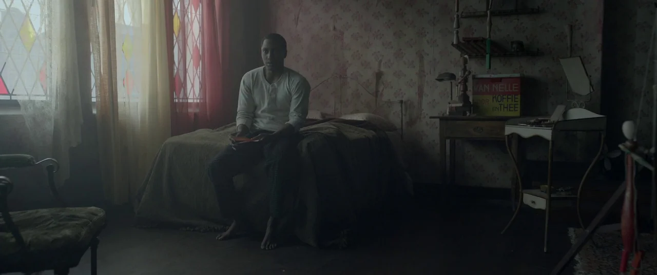 Amsterdam (2022) movie still: medium — A dimly lit room features floral wallpaper and a vintage wooden bedside table. A man sits motionless on the…