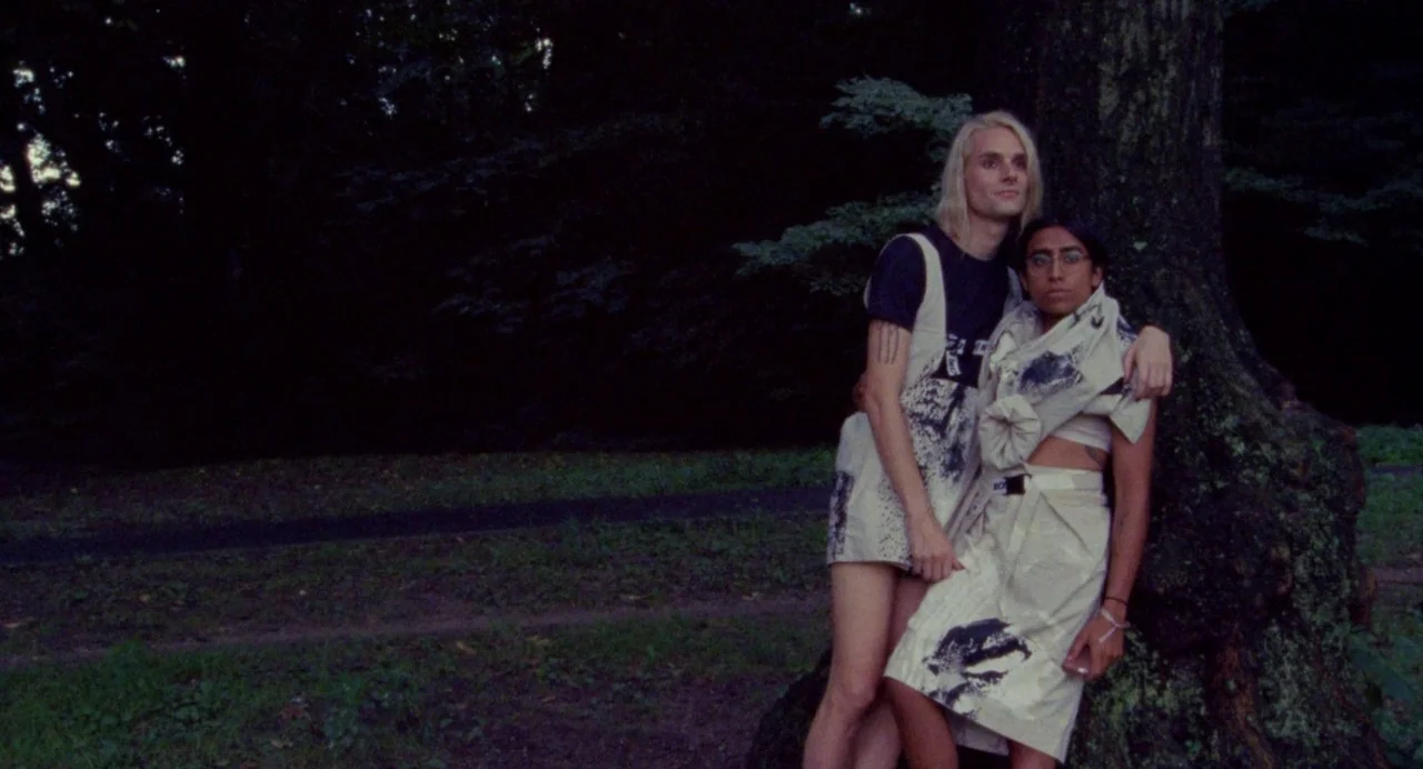 So Pretty (2019) movie still: fullbody — Two individuals stand closely together against a large tree in a dark wooded area. The background is a…