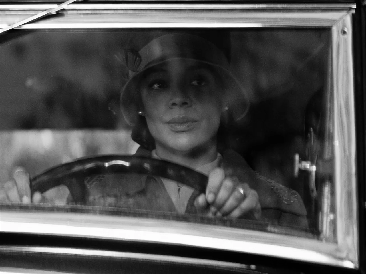 Passing (2021) movie still: closeup — A 1920s vehicle interior framed by the window frame and metal dashboard. A woman wearing a cloche hat grips…