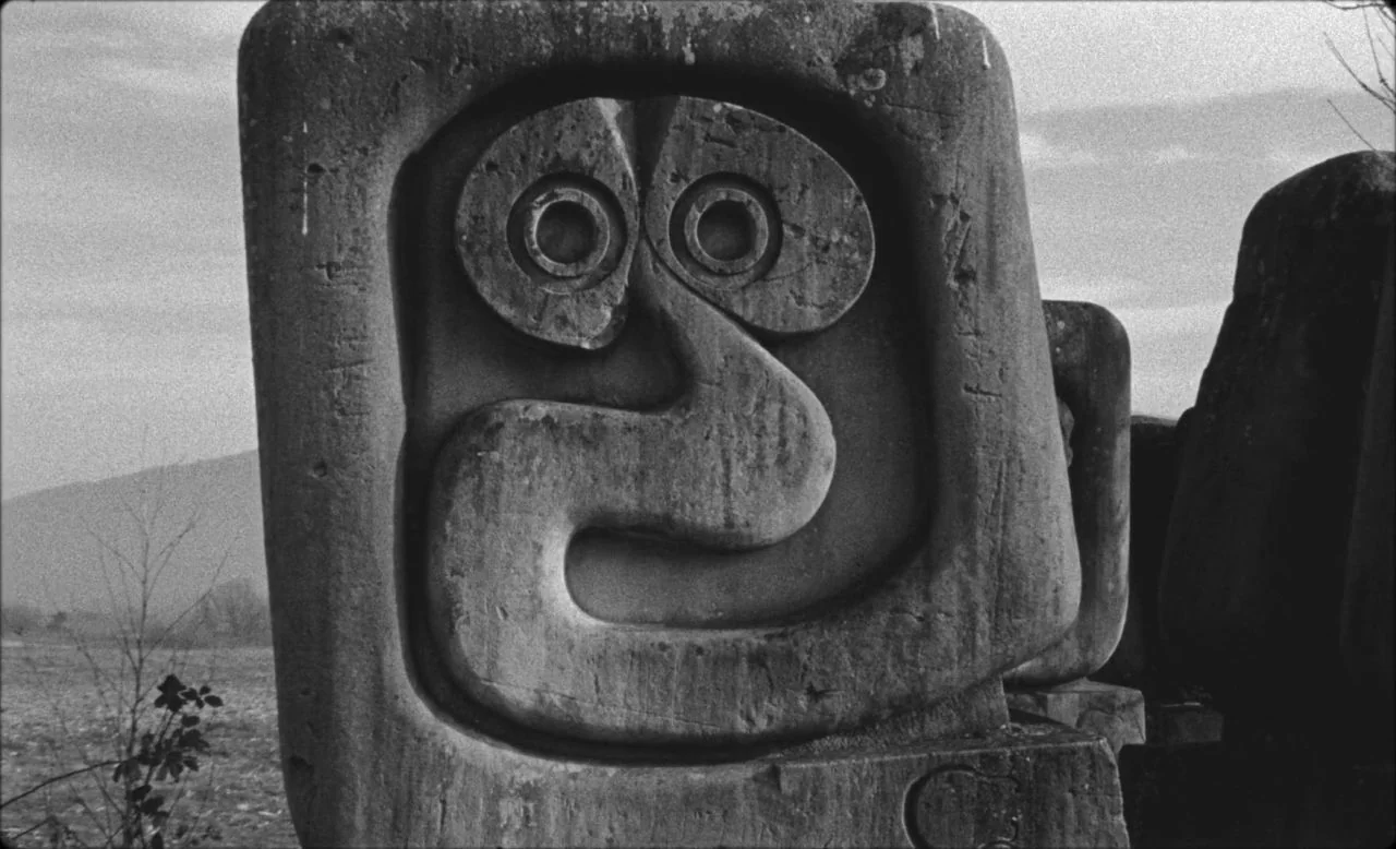 Last and First Men (2020) movie still: closeup — A monolithic stone block features a carved abstract face with prominent circular eyes. The…