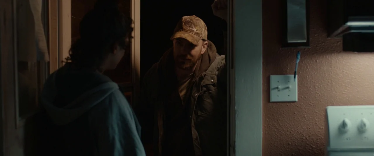 Little Woods (2018) movie still: medium — A dimly lit doorway separates a dark exterior from a warm interior space. A man wearing a camouflage cap…