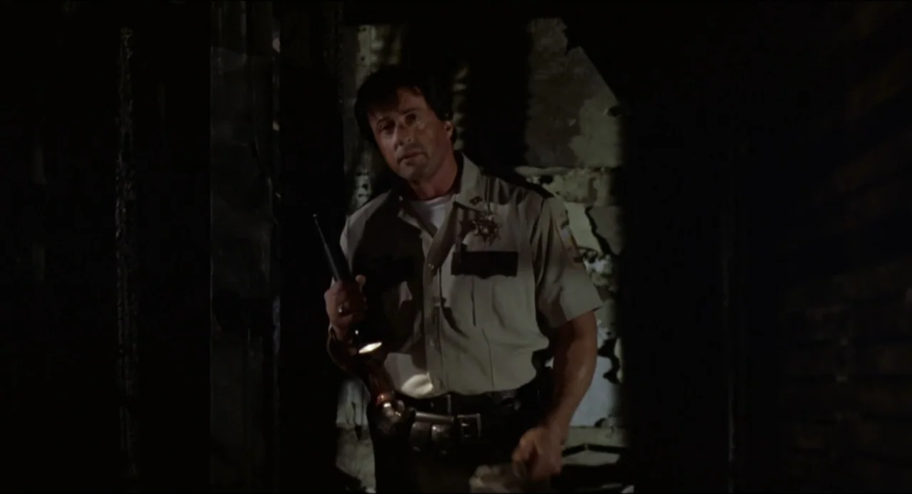 Cop Land (1997) movie still: medium — A dark, dilapidated indoor space with damaged plaster walls. A man in a law enforcement uniform walks through…