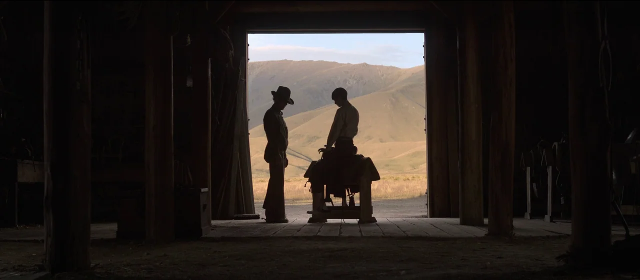 The Power of the Dog (2021) movie still: wide — A rustic barn interior overlooks a vast, arid mountain landscape in the distance. Two men stand in…