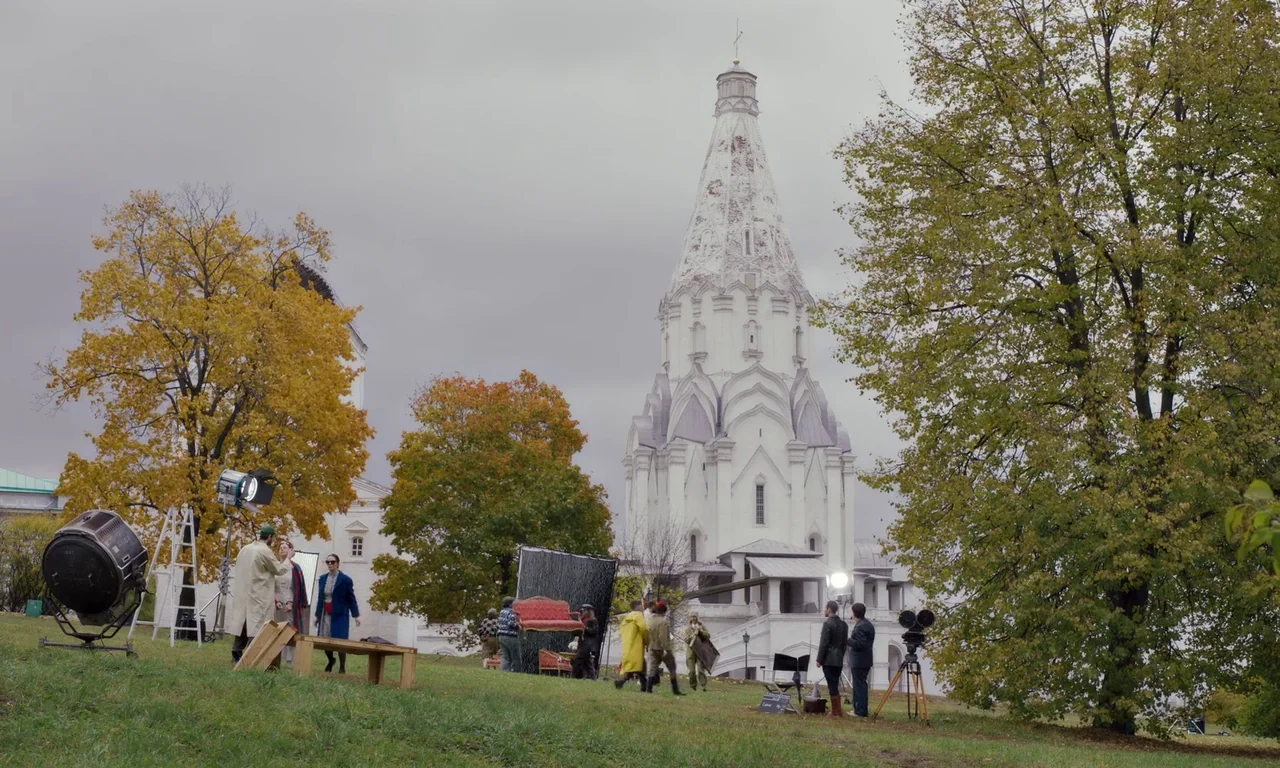 Bloodsuckers (2021) movie still: wide — A film crew works on a grassy slope with a large white stone church behind. Various cameras and light rigs…