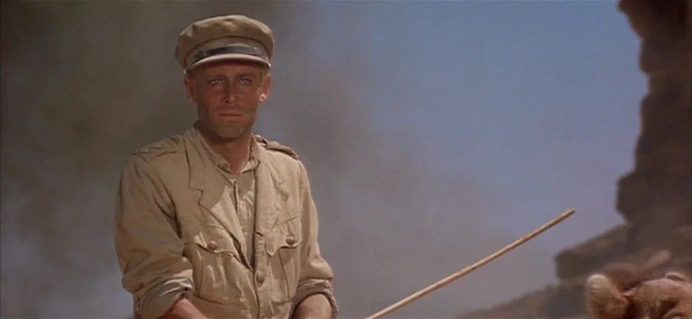 Lawrence Of Arabia movie still: medium — An arid, hazy desert landscape features a man standing in the foreground. He wears a tan military uniform…