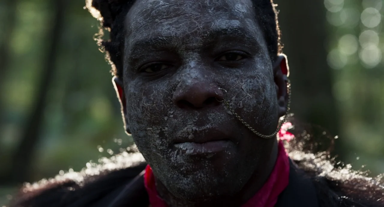 Zombi Child (2019) movie still: closeup — A person stands in a wooded area with light filtering through the surrounding trees. Their face is heavily…