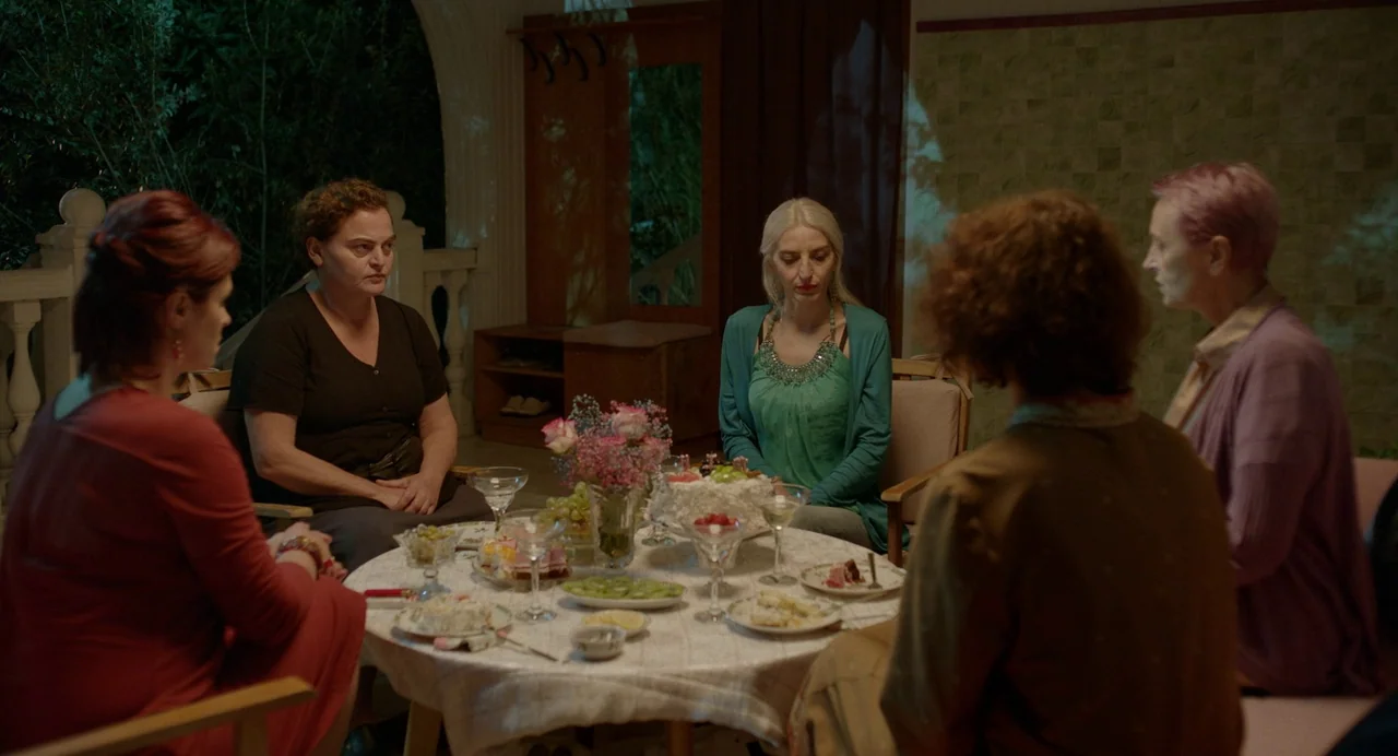 Blackbird Blackbird Blackberry (2023) movie still: wide — A group of five women sit around a circular table on a dark terrace. They are gathered for…