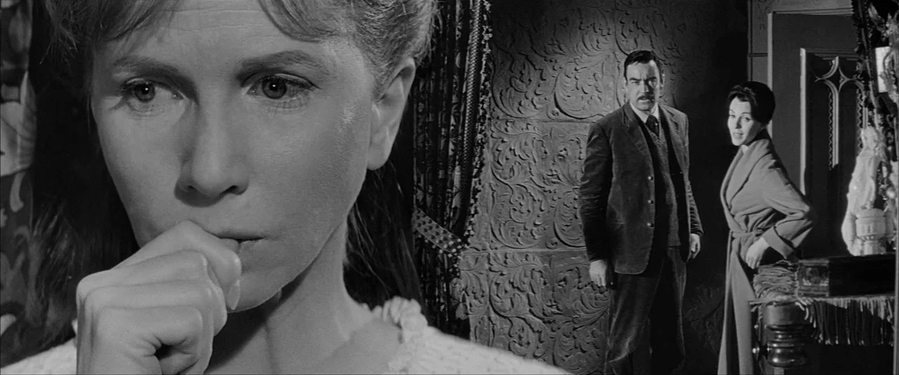 The Haunting (1963) movie still: closeup — A dimly lit Victorian interior features ornate embossed wallpaper and heavy draped fabric. A woman in the…