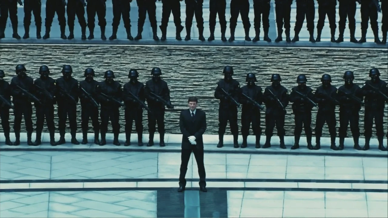Ultraviolet (2006) movie still: wide — A stone courtyard features a long wall with a row of armored guards behind it. A man in a dark suit stands…