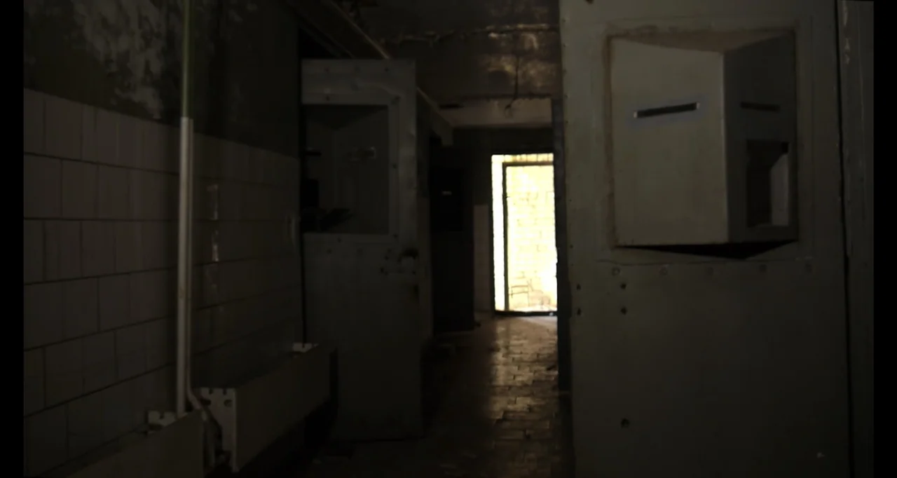 Chernobyl's Café (2016) movie still: wide — A dark, dilapidated hallway features metal cell doors with viewing ports along the wall. The space is…