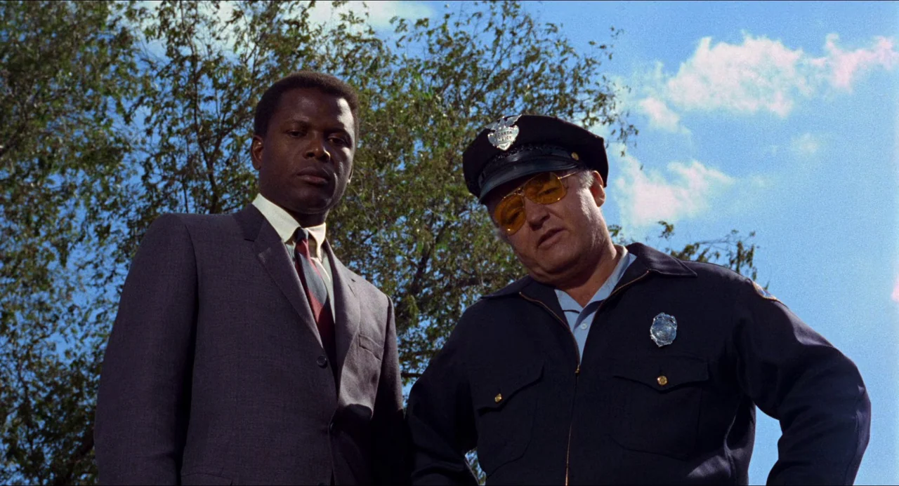 In the Heat of the Night (1967) movie still: medium — Two men stand outdoors under a bright blue sky with sparse tree branches above. The men gaze…