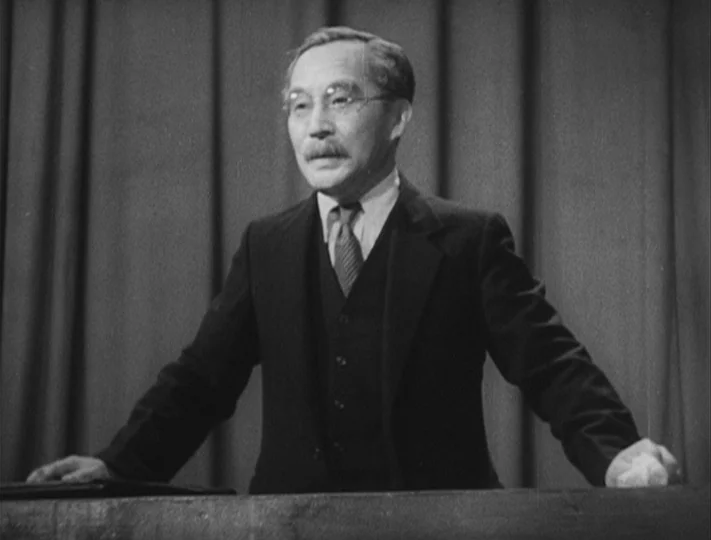 No Regrets for Our Youth (1946) movie still: medium — An elderly man stands at a lectern backed by heavy, draped curtains. He addresses an audience…