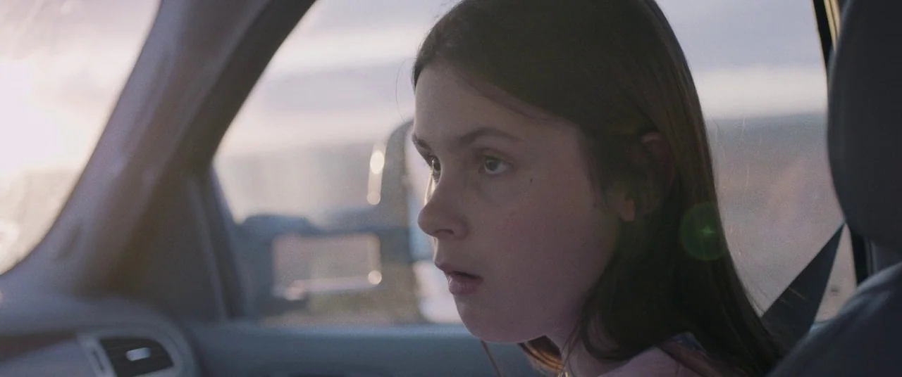 The Rider (2017) movie still: closeup — A child sits in the passenger seat of a car during the day. The girl looks out the window with her mouth…