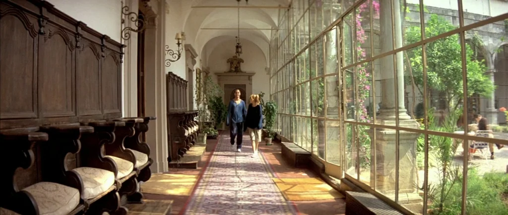 The Big Blue (1988) movie still: wide — A long, bright corridor features a row of wooden benches and arched ceilings. Two women walk forward along a…