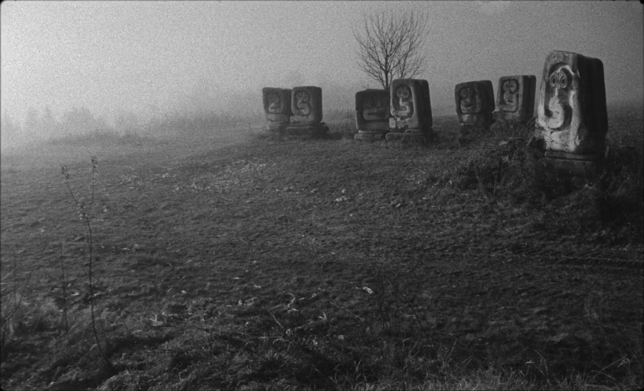 Last and First Men (2020) movie still: wide — A grassy, uneven field is blanketed in heavy morning fog. Several carved stone sculptures with…
